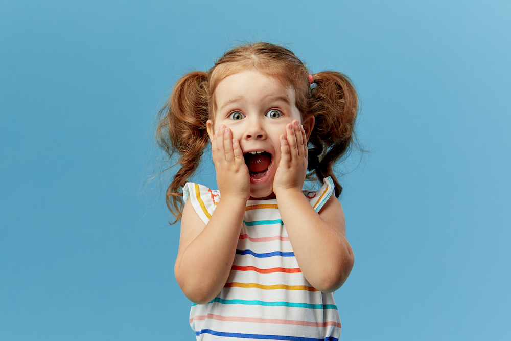 Portrait of surprised cute little toddler girl child standing isolated over blue background. Looking at camera. hands near open mouth Learn 7 crazy facts about braces from a Charleston SC orthodontist