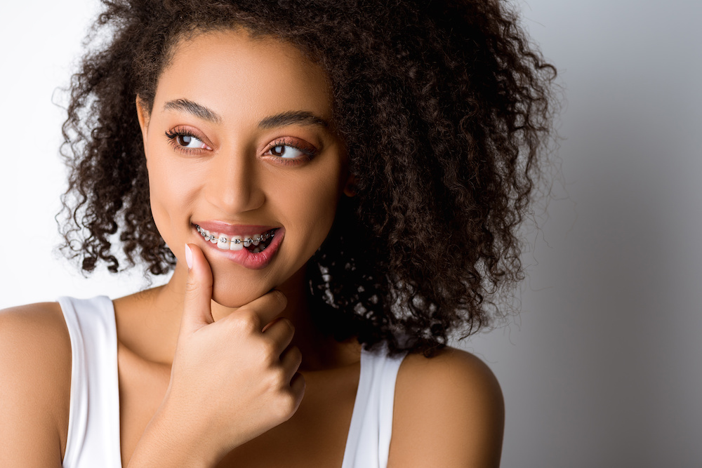 thoughtful smiling african american girl with dental braces touching lip, isolated on grey Things to remember when you have braces in Charleston SC