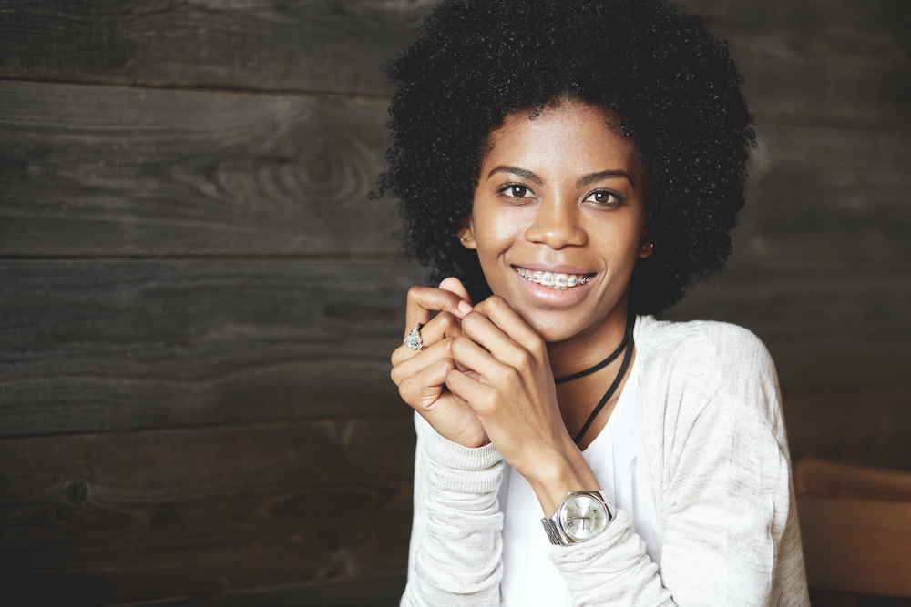 Close up shot of attractive happy cheerful dark-skinned young woman looking and smiling at the camera, showing white teeth with braces, holding hands at her face. Human face expressions and emotions Discover platinum braces in Charleston SC